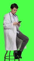 Side view Woman specialist using smartphone social media apps in studio, sitting on a chair against greenscreen backdrop. Physician waiting for someone and browses online webpages, texting. Camera A. video