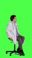 Side view Physician in white coat waiting for patients at consultations, feeling impatient sitting on a chair against greenscreen backdrop. Medic practitioner with stethoscope waits for people. Camera A. video