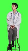 Front view General practitioner acting impatient sitting on a chair against greenscreen backdrop, waiting for patients to attend checkup appointments. Woman specialist in white coat waits for people. Camera A. video