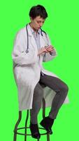 Side view General practitioner acting impatient sitting on a chair against greenscreen backdrop, waiting for patients to attend checkup appointments. Woman specialist in white coat waits for people. Camera A. video