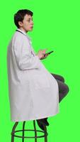 Profile Woman specialist using smartphone social media apps in studio, sitting on a chair against greenscreen backdrop. Physician waiting for someone and browses online webpages, texting. Camera A. video