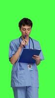 Front view Medical assistant writing checkup information on clipboard files, taking notes and making doctor appointments with checklist. Nurse wearing blue scrubs stands against greenscreen backdrop. Camera B. video