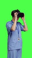 Side view Medical assistant uses interactive vr glasses to check disease treatment, looking at patient examination files with virtual reality headset and lens. Nurse against greenscreen backdrop. Camera B. video