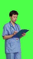 Side view Medical assistant writing checkup information on clipboard files, taking notes and making doctor appointments with checklist. Nurse wearing blue scrubs stands against greenscreen backdrop. Camera B. video