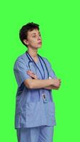 Side view Portrait of confident medical assistant posing with arms crossed in studio, standing against greenscreen backdrop. Successful nurse with healthcare expertise wearing blue scrubs uniform. Camera B. video