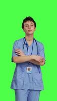 Front view Portrait of smiling medical assistant poses with arms crossed, showing confidence dressed in blue hospital scrubs. Successful nurse standing against greenscreen backdrop, health specialist. Camera B. video