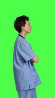 Profile Portrait of smiling medical assistant poses with arms crossed, showing confidence dressed in blue hospital scrubs. Successful nurse standing against greenscreen backdrop, health specialist. Camera B. video