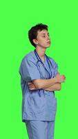 Side view Impatient nurse acting frustrated while she waits for patients at clinic, standing against greenscreen backdrop. Worried nurse with blue scrubs waiting for people to arrive at checkup. Camera B. video