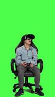 Front view Woman playing video games on console with controller and vr headset, sitting against greenscreen backdrop. Gamer having fun with online tournament, virtual reality interactive cyberspace. Camera A.