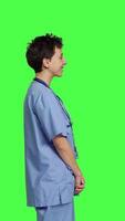 profil porträtt av medicinsk assistent leende och Framställ med förtroende, stående mot grönskärm bakgrund. sjuksköterska bär blå scrubs och stetoskop, känsla framgångsrik med hälsa expertis. kamera b. video