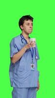 Side view Medical assistant drinking cup of coffee before starting exam appointments in the morning, enjoying caffeine refreshment against greenscreen backdrop. Nurse drinks beverage. Camera B. video