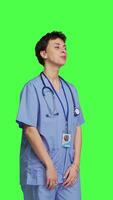 Side view Portrait of medical assistant smiling and posing with confidence, standing against greenscreen backdrop. Nurse wears blue scrubs and stethoscope, feeling successful with health expertise. Camera B. video