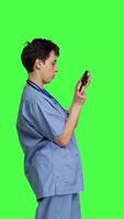 Profile Medical assistant playing mobile video games on smartphone app, taking a break from healthcare work. Nurse enjoys gaming tournament on phone, standing against greenscreen backdrop. Camera B.