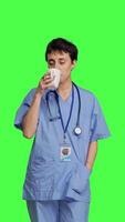 Front view Medical assistant drinking cup of coffee before starting exam appointments in the morning, enjoying caffeine refreshment against greenscreen backdrop. Nurse drinks beverage. Camera B. video