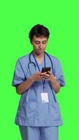 Front view Young nurse texting on smartphone against greenscreen backdrop, using mobile phone to check social media messages on work break. Medical assistant browsing online web pages. Camera B. video