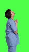 Profile Hospital nurse worshipping God by prayer against greenscreen backdrop, praying to jesus christ and having hope in christianity and spirituality. Medical assistant doing meditation. Camera B. video