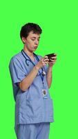 Side view Medical assistant playing mobile video games on smartphone app, taking a break from healthcare work. Nurse enjoys gaming tournament on phone, standing against greenscreen backdrop. Camera B.