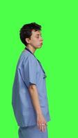 profiel ongeduldig verpleegster acteren gefrustreerd terwijl ze wacht voor patiënten Bij kliniek, staand tegen groene scherm achtergrond. bezorgd verpleegster met blauw scrubs aan het wachten voor mensen naar aankomen Bij controle. camera b. video