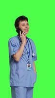 Side view Health specialist using smartphone to make important calls, standing against greenscreen backdrop. Nurse talking to patients for checkup exams on telephone line, calling people to confirm. Camera B. video