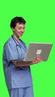 Side view Medical assistant browsing for healthcare treatments to help patients, using laptop against greenscreen backdrop. Young nurse navigates online websites an searches for health support. Camera B. video