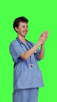 Side view Smiling medical assistant applauding someone in studio, celebrating success and cheering against greenscreen backdrop. Young nurse clapping hands and feeling happy about achievement. Camera B. video