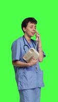 Side view Nurse answering landline phone call to make checkup appointments, talking to patients on telephone with cord against greenscreen backdrop. Health specialist in scrubs uses vintage phone. Camera B. video