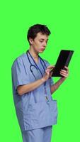 Side view Medical assistant browsing online webpages on tablet and texting, using social media apps to chat with people. Nurse navigates internet on gadget, standing against greenscreen backdrop. Camera B. video