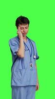 Side view Unwell nurse suffering from a headache against greenscreen backdrop, wearing scrubs and feeling overworked at hospital. Medical assistant having a painful migraine, stress and burnout. Camera B. video
