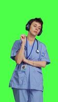 Front view Nurse having fun listening to music on wireless headphones, wearing scrubs and dancing around against greenscreen backdrop. Medical assistant showing groovy funky dance moves. Camera B. video