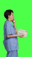 Profile Nurse answering landline phone call to make checkup appointments, talking to patients on telephone with cord against greenscreen backdrop. Health specialist in scrubs uses vintage phone. Camera B. video