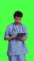 Front view Medical assistant browsing online webpages on tablet and texting, using social media apps to chat with people. Nurse navigates internet on gadget, standing against greenscreen backdrop. Camera B. video