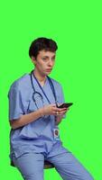 Side view Nurse browsing online websites on smartphone app in studio, sitting on a chair against greenscreen backdrop. Medical specialist in scrubs using mobile phone and social media texting. Camera B. video