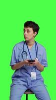 Front view Nurse browsing online websites on smartphone app in studio, sitting on a chair against greenscreen backdrop. Medical specialist in scrubs using mobile phone and social media texting. Camera B. video