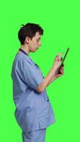 Profile Medical assistant browsing online webpages on tablet and texting, using social media apps to chat with people. Nurse navigates internet on gadget, standing against greenscreen backdrop. Camera B. video