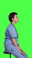 Profile Medical assistant waiting for someone while she sits on chair, feeling impatient and waits for patients to attend checkup appointments. Nurse in scrubs against greenscreen backdrop. Camera B. video