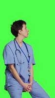 Side view Medical assistant waiting for someone while she sits on chair, feeling impatient and waits for patients to attend checkup appointments. Nurse in scrubs against greenscreen backdrop. Camera B. video