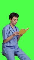 Side view Young nurse studying for the physician exam by reading book in studio, sitting on chair against greenscreen. Specialist enjoys healthcare industry literature, preparing to become a doctor. Camera B. video