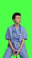 Front view Medical assistant waiting for someone while she sits on chair, feeling impatient and waits for patients to attend checkup appointments. Nurse in scrubs against greenscreen backdrop. Camera B. video