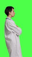 Profile Woman medic posing with arms crossed in a white coat, feeling confident and successful with her medical career and expertise. Doctor having a stethoscope standing against greenscreen. Camera B. video