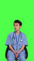 Front view Medical assistant is waiting for patients to attend exam appointments, feeling impatient while she sits in a chair. Nurse with stethoscope and scrubs waits for people against greenscreen. Camera B. video