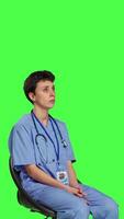 Side view Medical assistant is waiting for patients to attend exam appointments, feeling impatient while she sits in a chair. Nurse with stethoscope and scrubs waits for people against greenscreen. Camera B. video