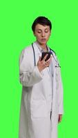 Side view Woman specialist looking at phone watch and waiting for patients to arrive at exam appointments, greenscreen backdrop in studio. General practitioner in white coat acting impatient. Camera B. video