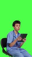 Side view Medical assistant browsing internet on tablet while she sits down in a chair, killing time by scrolling on social media apps and texting friends. Nurse in scrubs using modern gadget. Camera B. video