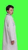 Profile Physician saying no and showing negative reaction symbol in studio, wearing a white coat and standing against greenscreen backdrop. Woman medic feeling displeased, rejection sign. Camera B. video