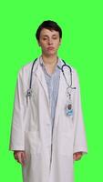 Front view Physician saying no and showing negative reaction symbol in studio, wearing a white coat and standing against greenscreen backdrop. Woman medic feeling displeased, rejection sign. Camera B. video