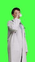 Side view Woman physician drinking a cup of coffee against greenscreen backdrop, taking a break from medical work. Specialist serving caffeine refreshment in studio, wearing a white coat. Camera B. video