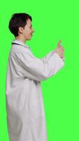 Profile Cheerful medic doing thumbs up symbol against greenscreen backdrop, expresses positivity with like sign. General practitioner with coat giving approval and being satisfied with success. Camera B. video