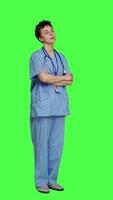 kant visie portret van glimlachen medisch assistent poses met armen gekruist, tonen vertrouwen gekleed in blauw ziekenhuis schrobt. geslaagd verpleegster staand tegen groene scherm achtergrond, Gezondheid specialist. camera a. video