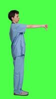 Profile Displeased medical assistant shows thumbs down in studio, presenting her disapproval and disagreement with dislike symbol against greenscreen. Nurse feeling unhappy with rejection sign. Camera A. video