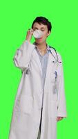 Front view Woman physician drinking a cup of coffee against greenscreen backdrop, taking a break from medical work. Specialist serving caffeine refreshment in studio, wearing a white coat. Camera B. video